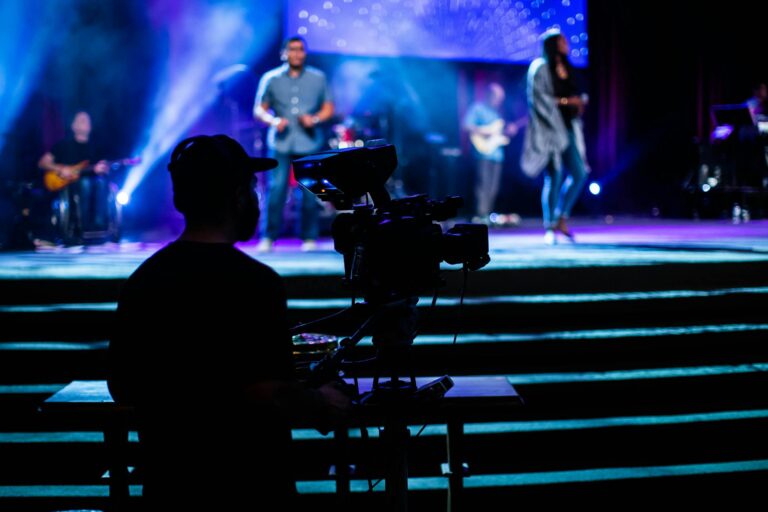 Silhouette of a cameraman filming a vibrant live concert with musicians and colorful stage lights.